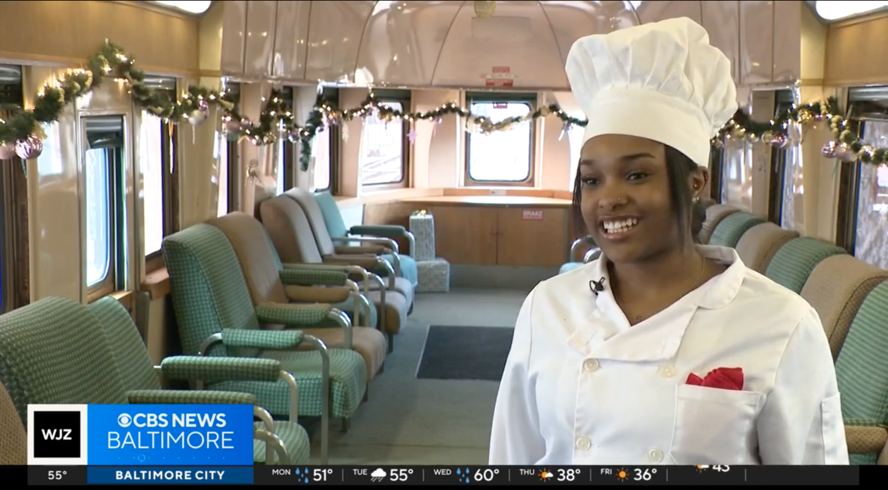 This is an image of a woman standing on a train wearing a chef's jacket and chef's hat.